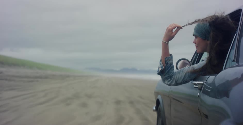 Slow motion of a woman's hair blowing in the wind as she rides in a vintage car driving along a sandy beach