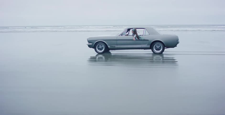 A classic grey coupe cruises along a wide, reflective beach as a woman leans out the window to feel the ocean air