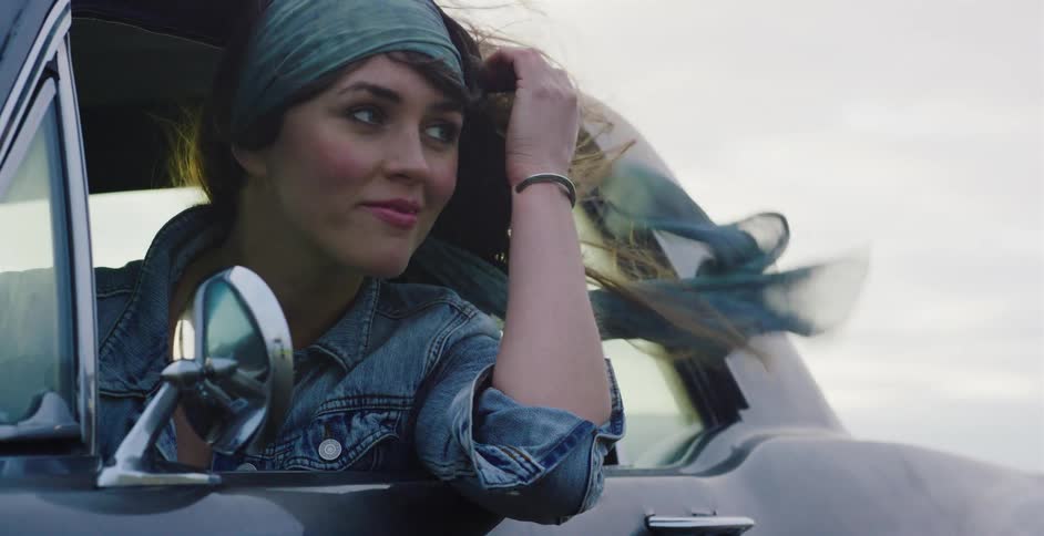A carefree young woman smiles as she leans out of a classic car window, her hair and scarf blowing in the wind