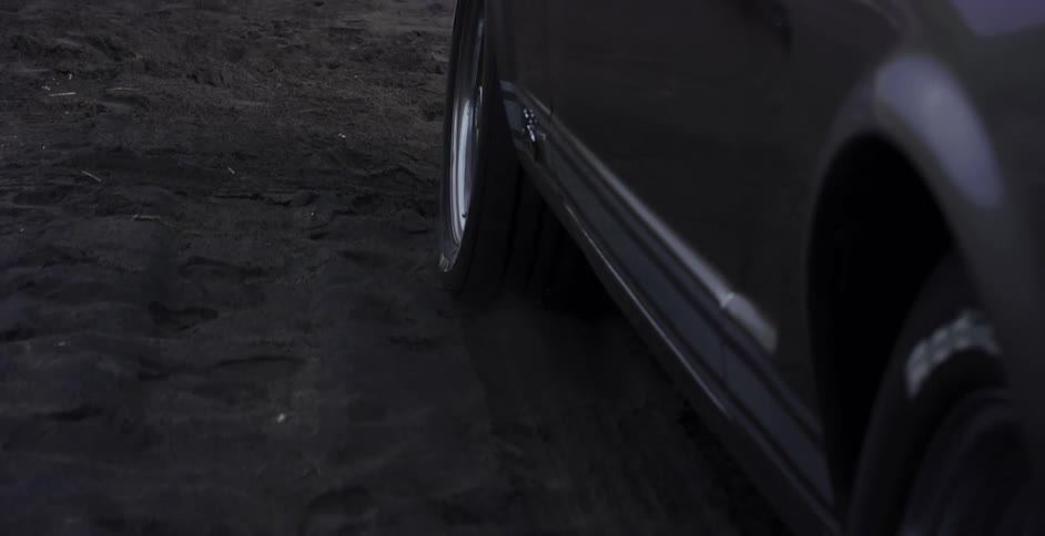 Close-up of a classic car's tire spinning and doing a burnout in black sand in slow motion