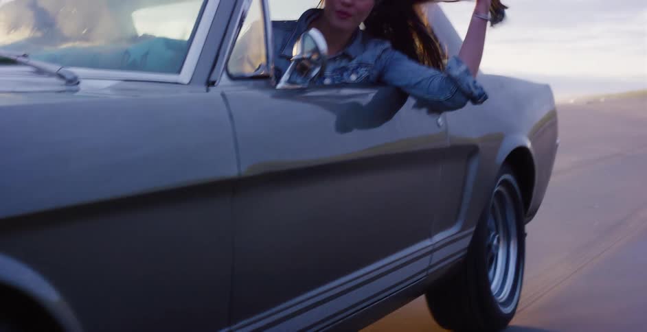 A young woman leans out the window of a classic Ford Mustang, playing with her hair as she drives along a wet sand beach at dusk