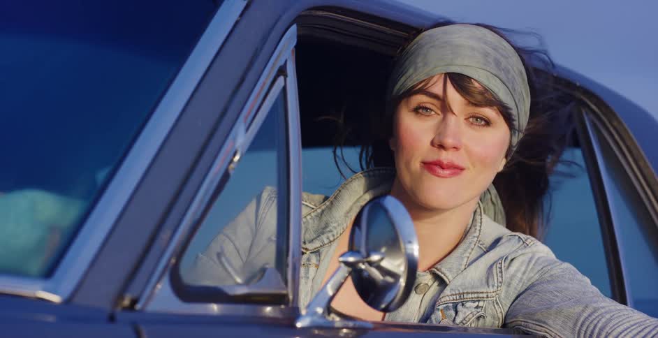 A beautiful woman with wind in her hair smiles as she leans out the window of her vintage car on a scenic drive at sunset