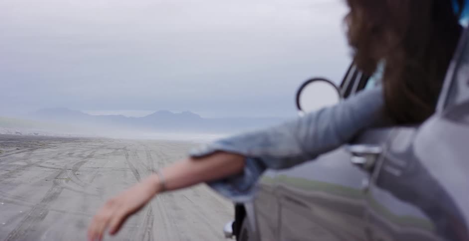Slow motion over-the-shoulder shot of a woman's arm hanging out of a classic car window as she drives along a vast, foggy beach