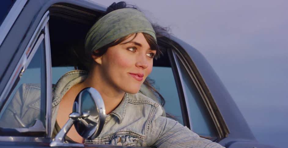 A beautiful young woman with windblown hair smiles as she enjoys a ride in a classic car during the golden hour