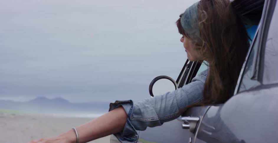 Slow motion shot of a young woman with windblown hair smiling as she leans out of a car window on a vast, cloudy beach