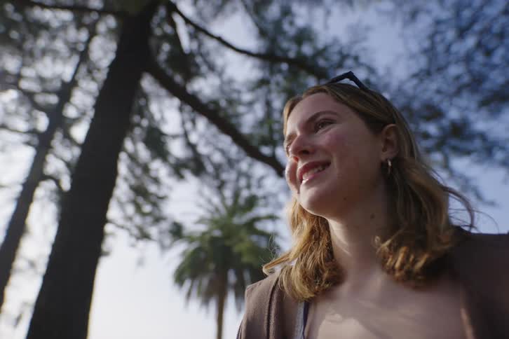 A smiling young woman looks up with joy, bathed in the sunlight filtering through tall pine and palm trees