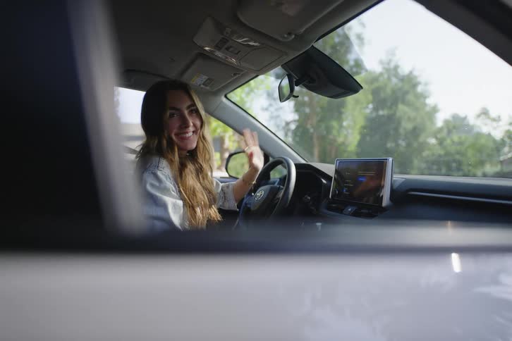Friendly young woman smiles and waves hello from the driver's seat of her car