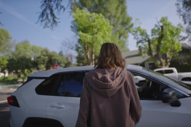 A woman in a hoodie approaches a white SUV and opens the back passenger door to join her friend in the driver's seat on a sunny day