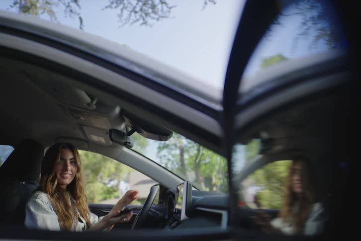 A cheerful young woman sitting in the driver's seat of her parked car looks up from her phone and smiles warmly at the camera