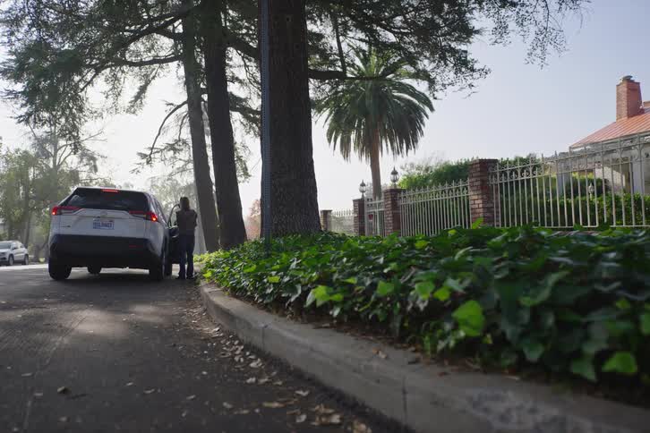 A woman gets into her white SUV parked on a quiet, tree-lined residential street during the day