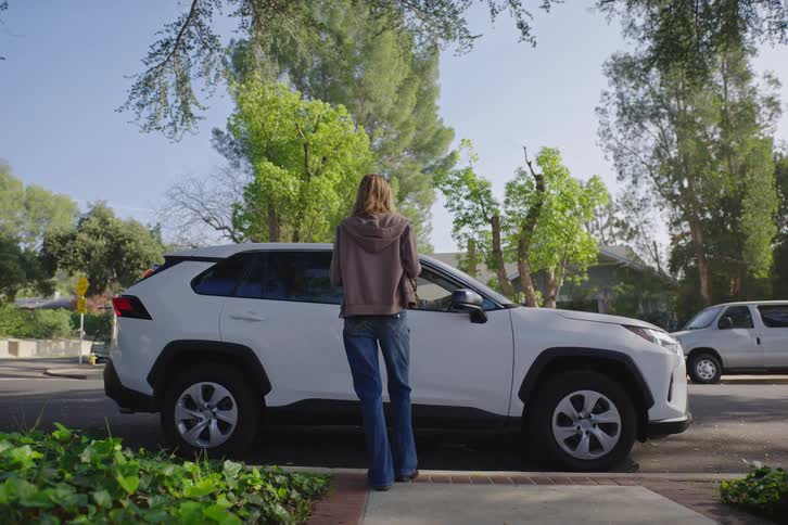 A person in a hoodie and jeans stands next to a white SUV parked on a suburban street