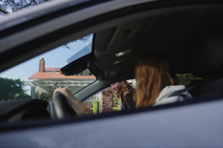 A young woman smiles as her friend gets into the passenger seat of her car parked outside a suburban home