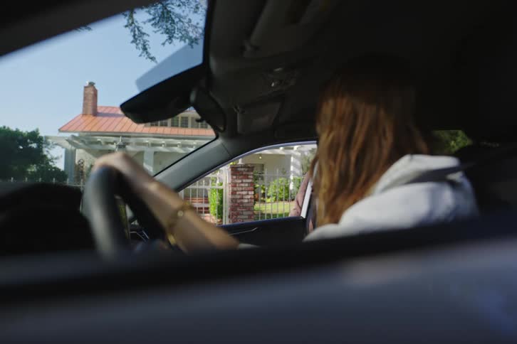 A young woman watches from the driver's seat as her friend gets into the passenger side of the car
