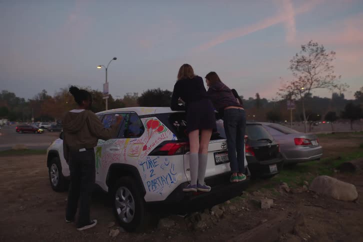 A diverse group of teenage friends celebrate together by drawing all over a white car in a parking lot at dusk