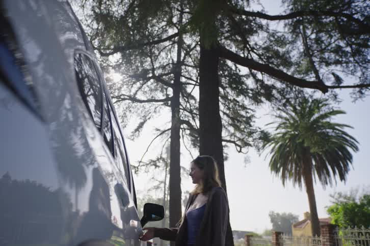 A smiling young woman waves as she walks to her car, unlocks the door, and gets in on a sunny suburban street