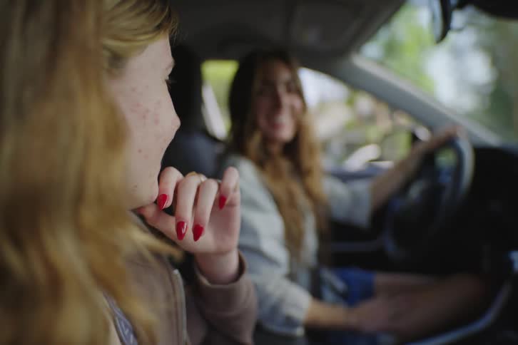 A teenage girl smiles at her friend in the passenger seat while they talk in a parked car