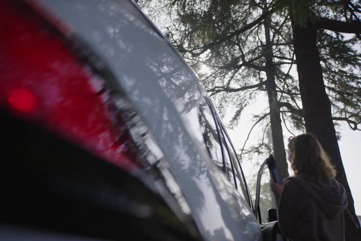 From a low angle, a woman approaches the passenger side of a car and gets in