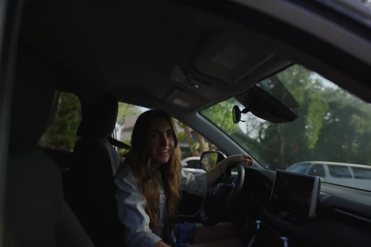 A young woman gets into the driver's seat of her car and smiles while chatting with a friend on a sunny suburban street