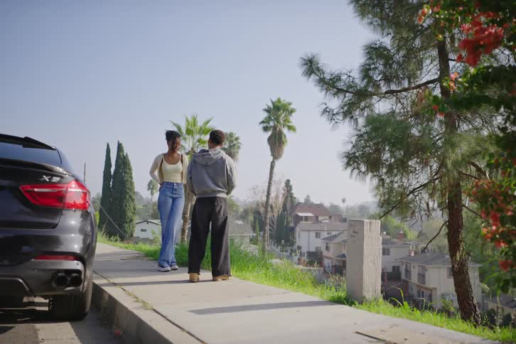 A young man and woman stand on a sunny sidewalk on a hillside street, engaged in a friendly conversation