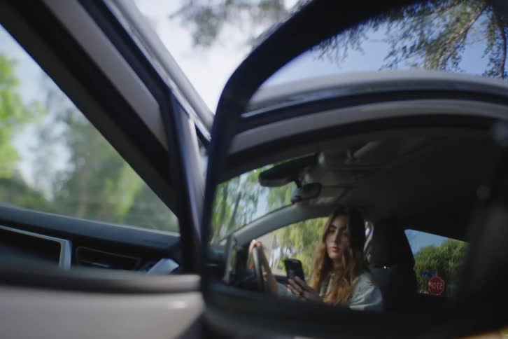 A smiling woman looks up from her smartphone, seen in the reflection of a car's side-view mirror