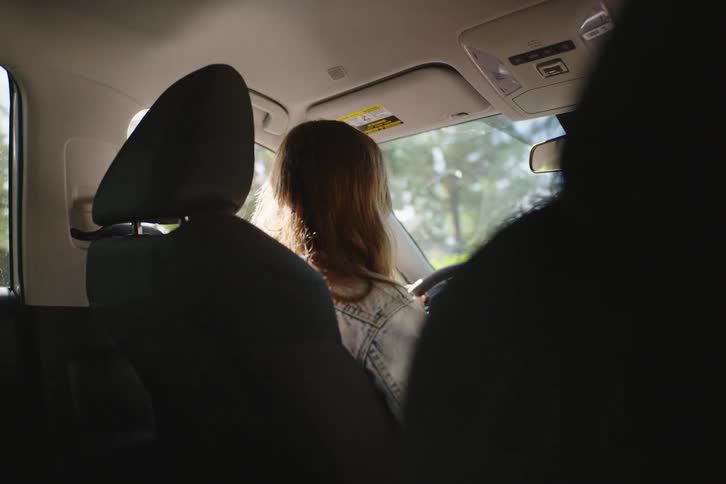From the backseat, a woman with long brown hair drives down a sunlit road