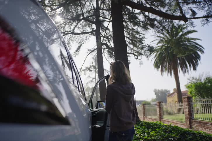 A woman walks up to her parked car on a sunny suburban street, opens the driver's side door, and gets in