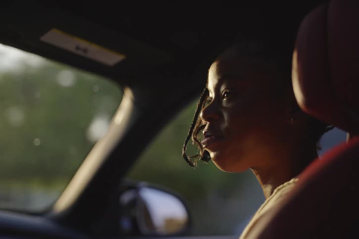 A thoughtful young woman with braids sits in the passenger seat enjoying a car ride at sunset