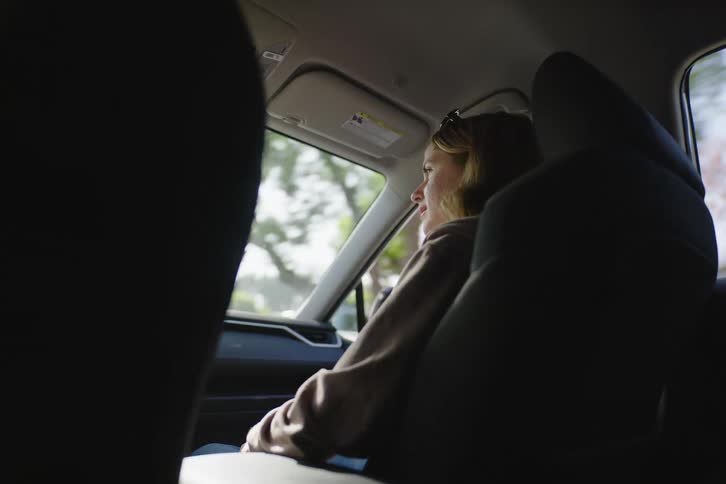 A pensive young woman looks out the window while riding in the back seat of a car on a sunny day