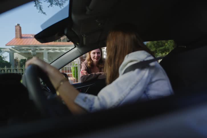 A young woman in the driver's seat of her car talks to her friend leaning through the open passenger window on a suburban street