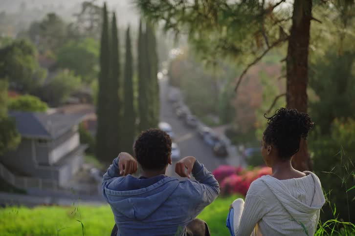 From behind, a young couple sits together on a lush green hill, sharing a peaceful conversation while overlooking a quiet suburban street