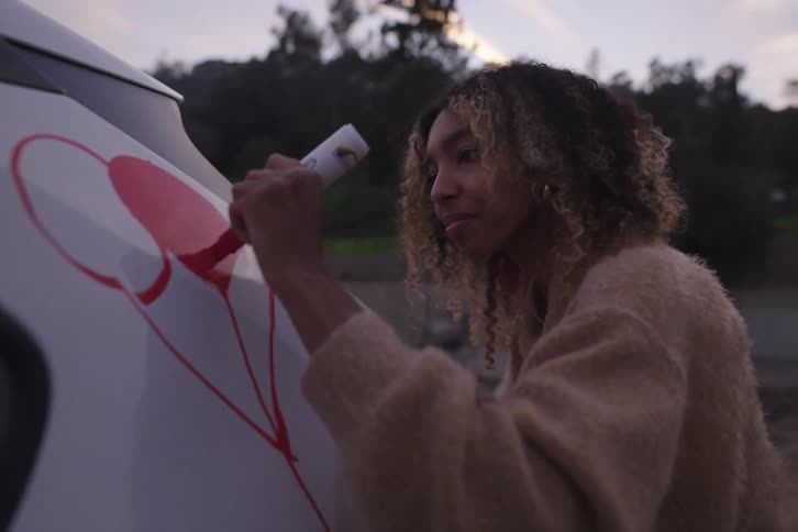 A happy young woman smiles as she creatively draws red heart-shaped balloons on the side of a white car at twilight