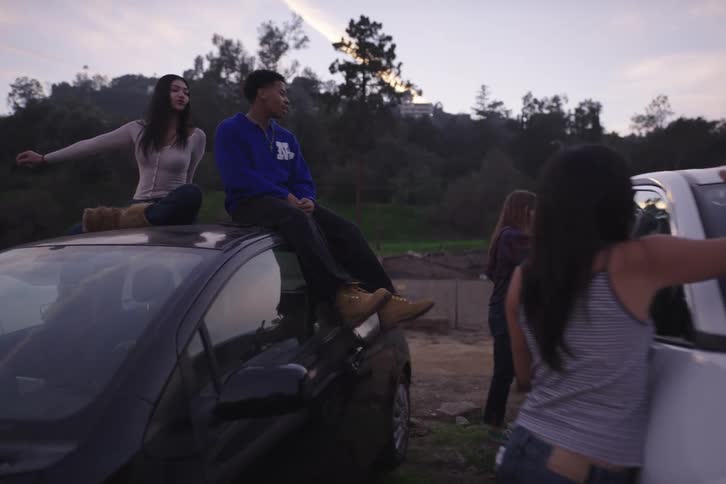A group of diverse friends hang out at dusk, sitting on cars and drawing on the windshield