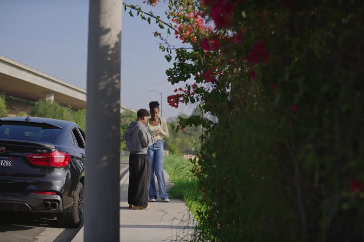 Two women stand on a sunny sidewalk next to a black SUV and flowering bushes, looking at a smartphone together