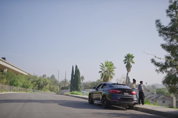 A couple stands beside their parked SUV on a quiet hillside road, talking and taking in the view on a sunny day