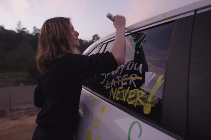 A student uses colorful markers to write humorous goodbye messages on a car for her graduating friends at sunset