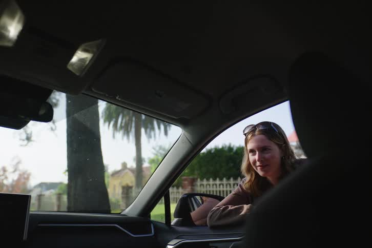 A young woman leans into a car window to have a friendly conversation with someone inside, as seen from the passenger seat