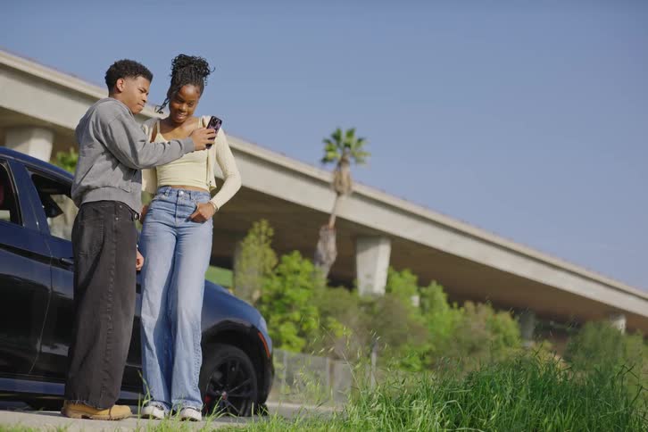 A young Black couple smiles while looking at a smartphone together, standing next to their car by an urban overpass on a sunny day