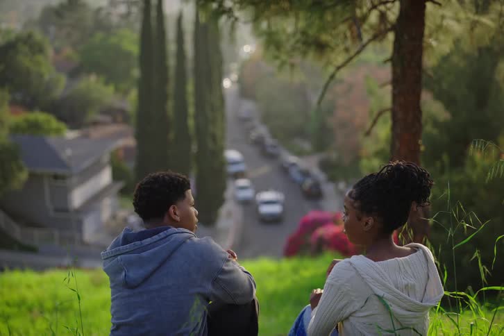 A young Black man and woman sit together on a grassy hill, having a conversation while overlooking a peaceful suburban street