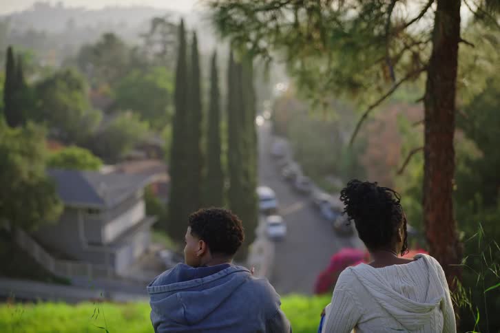 A young man and woman sit together on a grassy hill, engaged in a conversation while overlooking a peaceful suburban street