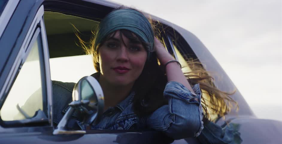 A beautiful young woman in a vintage car smiles at the camera as the wind blows through her hair in slow motion