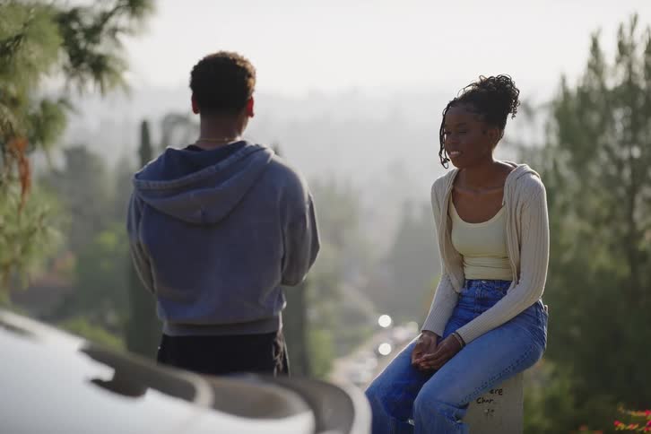 A young Black couple has a conversation on a scenic hillside overlook with a hazy city view in the background