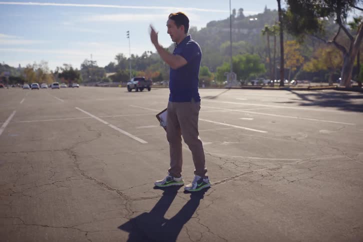 A man with a clipboard walks through a sunny parking lot, gesturing to someone off-camera and then taking notes