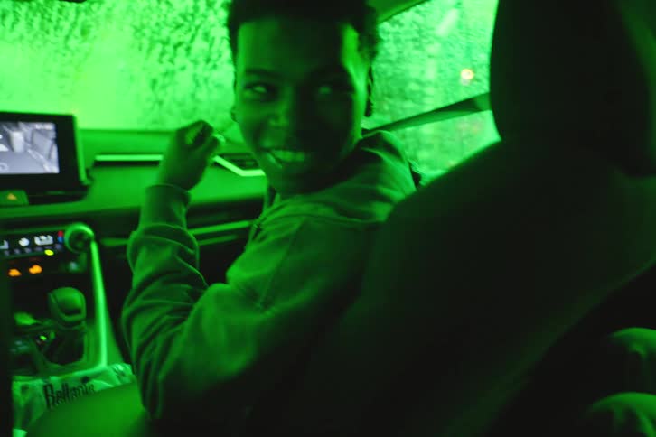A group of young people smiles and laughs while riding in a car through a tunnel of red and green neon lights at night