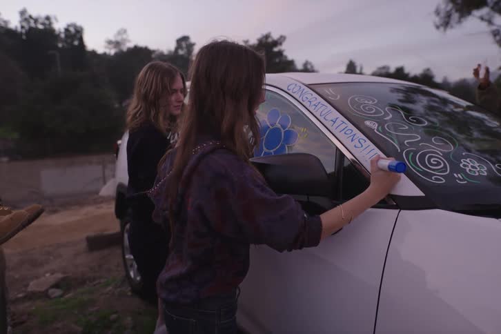 Friends gather at dusk to joyfully decorate a car with colorful markers, writing 'Congratulations' on the window to celebrate a special occasion