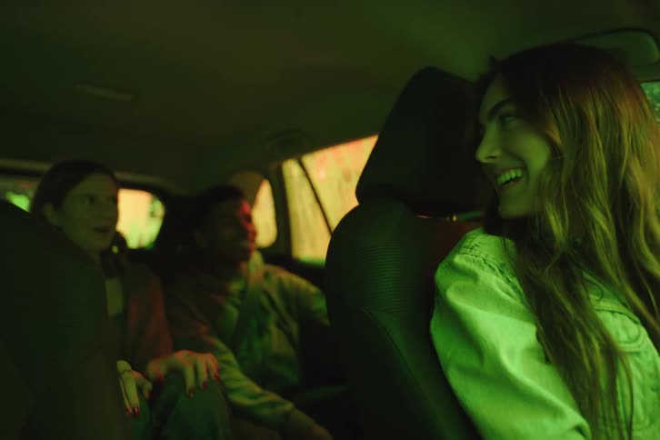 A young woman and her friends smile and laugh while going through a colorful, neon-lit automatic car wash at night