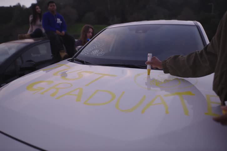 A group of happy friends celebrate graduation by writing 'Just Graduate' on the hood of a car with markers at dusk
