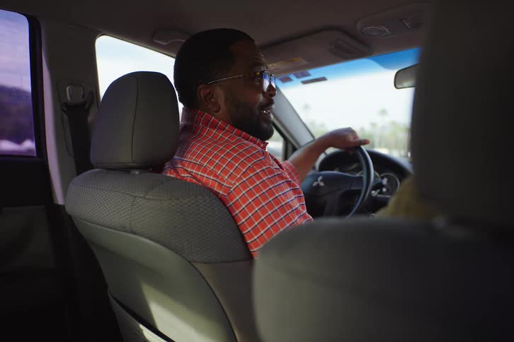 View from the back seat of a man in a red plaid shirt having an animated conversation with a passenger in his car