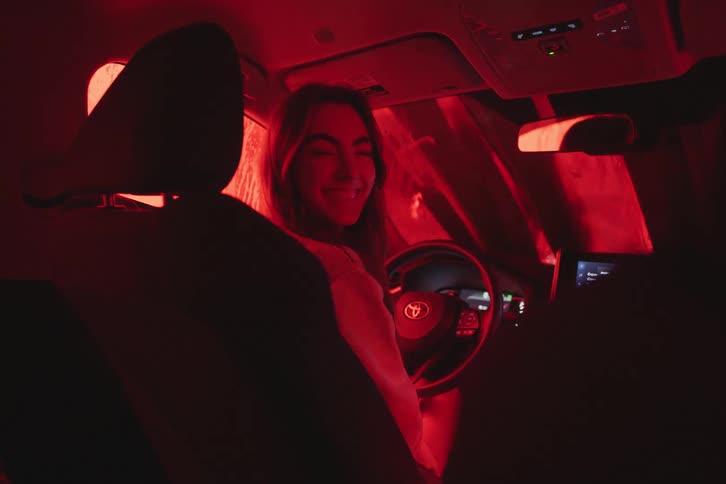 A woman smiles at her passenger as they drive through a colorful car wash at night, illuminated by changing green and red lights