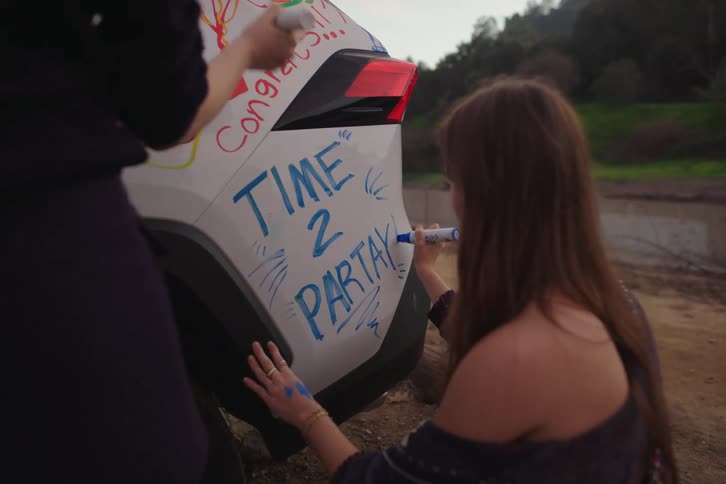 Two friends celebrate by writing celebratory messages with markers all over a white car
