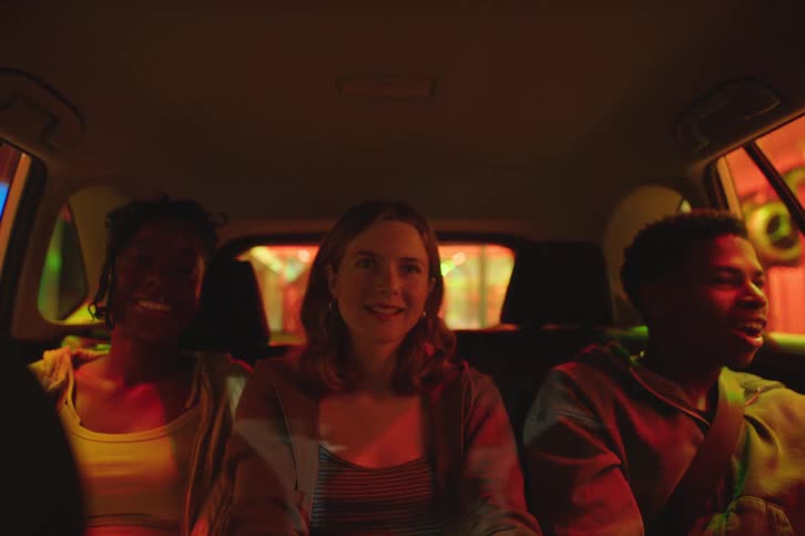 Three happy, diverse friends laugh and talk in the back of a car at night as colorful city lights flash across their faces